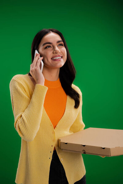 happy young woman calling on smartphone while standing with pizza box isolated on green