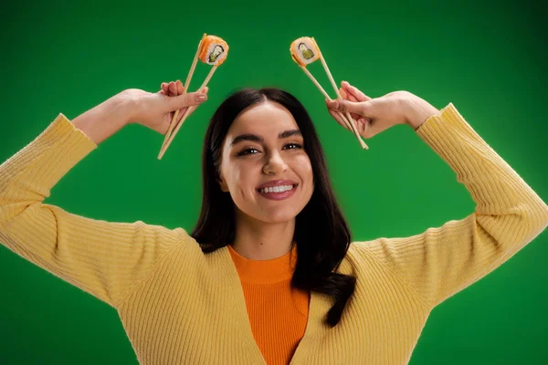 stock image cheerful woman in yellow jumper holding fresh sushi rolls with chopsticks isolated on green