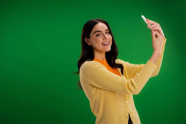 stock image cheerful woman looking at camera while taking selfie on cellphone isolated on green