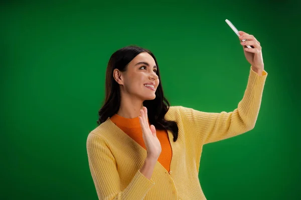 happy and stylish woman waving hand during video call on smartphone isolated on green