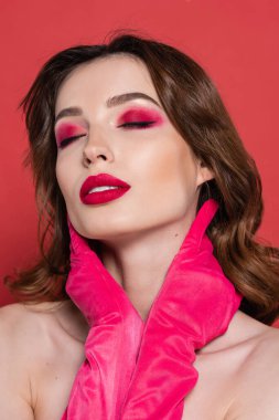 portrait of young woman with magenta color eye shadow touching neck isolated on pink 