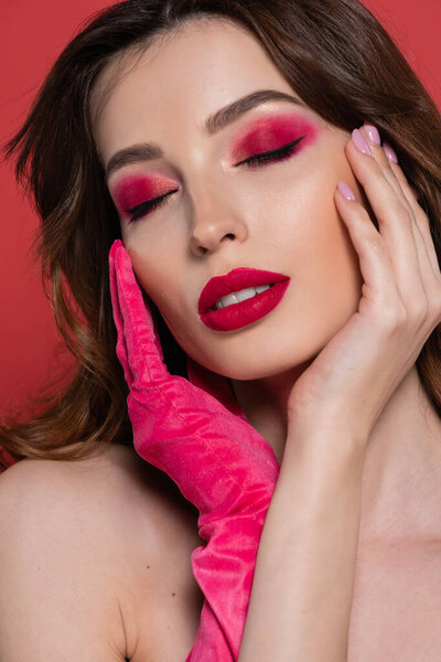 portrait of young woman with magenta color eye shadow gently touching face with hands in glove isolated on pink 