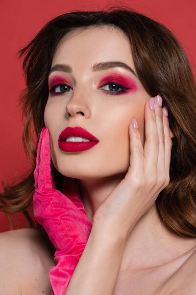 portrait of young woman with magenta color glove looking at camera while touching face isolated on pink 
