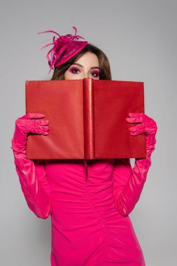 young and brunette woman in magenta color gloves and hat with feather covering face with book isolated on grey  clipart