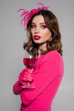 portrait of brunette woman in elegant hat and magenta color dress holding glass with alcohol drink isolated on grey 