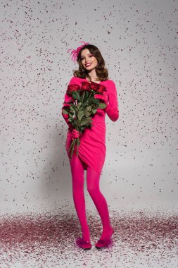 full length of happy woman in hat and magenta color dress holding red roses on grey with falling confetti 