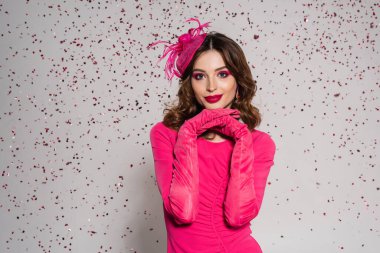 cheerful woman in elegant hat and magenta color dress posing on grey background with falling confetti 