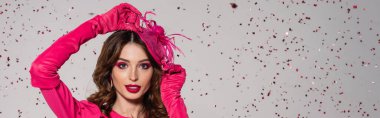 young brunette woman in magenta color gloves adjusting elegant hat near falling confetti on grey, banner 