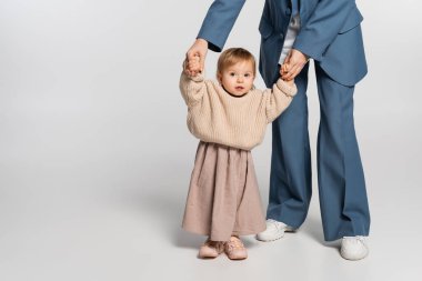 mother in blue suit standing and holding hands of toddler girl on grey  clipart