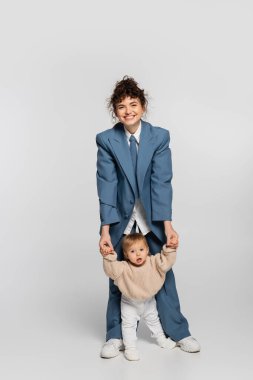happy mother in blue suit standing and holding hands of toddler girl on grey