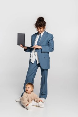 happy mother in blue suit standing with laptop while looking at toddler daughter on grey