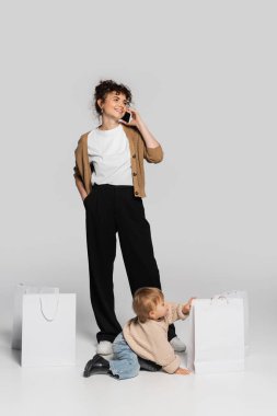 full length of happy woman in casual clothes talking on smartphone near toddler baby and shopping bags on grey 