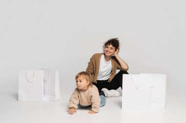 full length of cheerful mother in casual clothes sitting with toddler baby near shopping bags on grey 