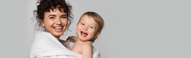 happy mother and cheerful toddler daughter covered in duvet isolated on grey, banner 