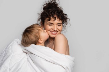 toddler baby girl kissing cheek of pleased mother covered in white duvet isolated on grey 