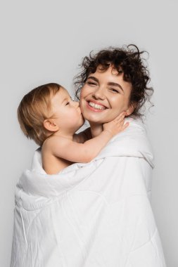 toddler baby girl kissing cheek of happy mother covered in white duvet isolated on grey 