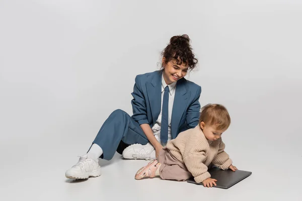 happy businesswoman in blue suit sitting with toddler daughter near laptop on grey