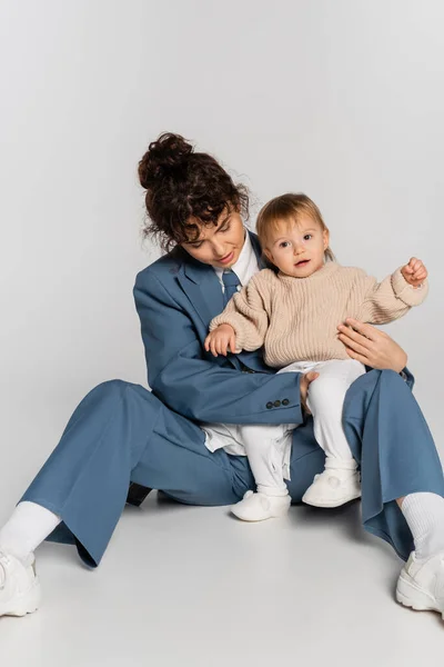 curly businesswoman in blue suit sitting with toddler daughter on grey background