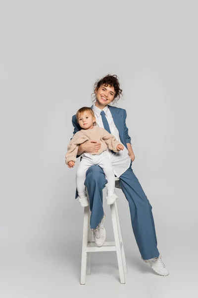 happy businesswoman in blue suit sitting with toddler daughter on high chair on grey
