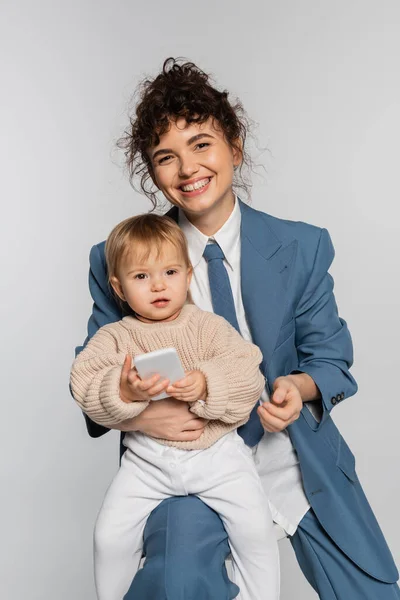 happy businesswoman in blue suit sitting on chair while toddler girl holding smartphone isolated on grey