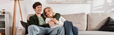 cheerful and curly man hugging happy blonde girlfriend in living room, banner 