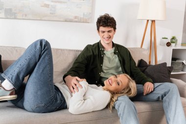 happy blonde woman lying on sofa near cheerful boyfriend  