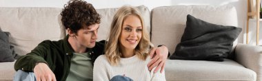 curly man hugging pretty blonde woman smiling near sofa in living room, banner 
