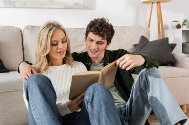 young couple smiling while reading book together in living room 