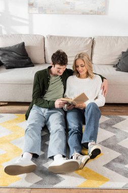 full length of cheerful couple smiling while reading book together in living room 