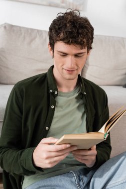 intelligent man with curly hair reading book in living room  clipart