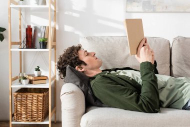 side view of young man with curly hair reading book while lying on couch in living room 