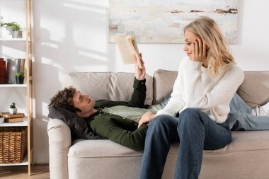 young man with curly hair reading book while lying on couch near smiling girlfriend in living room 