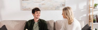 young and cheerful couple looking at each other in living room, banner 