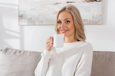dreamy young woman in white sweater holding cup of coffee and smiling at home 