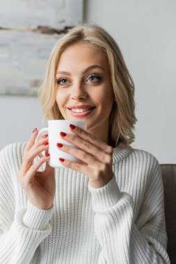 blonde young woman in white sweater holding cup of coffee and smiling at home 