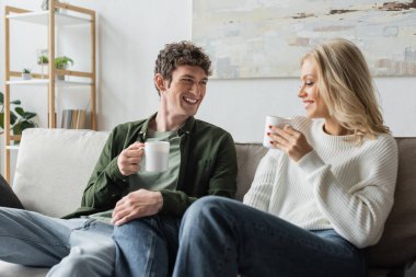 cheerful young man and blonde woman holding cups with coffee in living room 