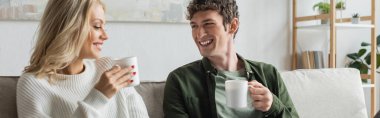 cheerful young man and blonde woman holding cups with coffee in living room, banner 