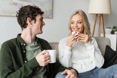 happy young man and blonde woman in sweater holding cups with coffee in living room 