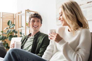 cheerful young man looking at blonde woman in sweater while holding cup with coffee in living room 