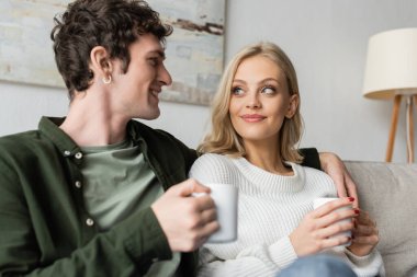 curly young man hugging blonde woman in sweater while holding cup with coffee in living room 