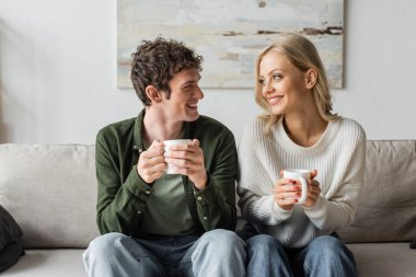 cheerful young man and blonde woman holding cups with coffee while looking at each other in living room 
