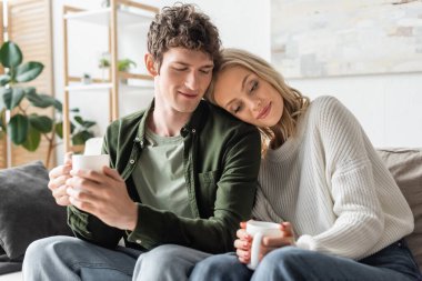 blonde woman in sweater holding cup with coffee and leaning on curly boyfriend in living room 