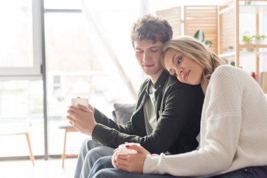 blonde woman in sweater holding cup of coffee and leaning on boyfriend in living room  clipart