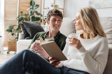 blonde woman in white sweater holding cup of coffee and reading book near cheerful boyfriend in living room 