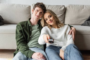 happy blonde woman clicking channels with remote controller near boyfriend in living room 