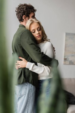 curly man hugging sensual blonde girlfriend near plant on blurred foreground 