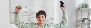 KYIV, UKRAINE - OCTOBER 24, 2022: excited young man in sweatshirt holding joystick in living room, banner 