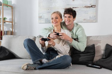 KYIV, UKRAINE - OCTOBER 24, 2022: cheerful young couple holding joysticks while playing video game on couch 