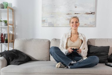 KYIV, UKRAINE - OCTOBER 24, 2022: cheerful young woman holding joystick while playing video game on couch 