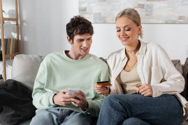 cheerful woman holding credit card near boyfriend with smartphone doing online shopping 
